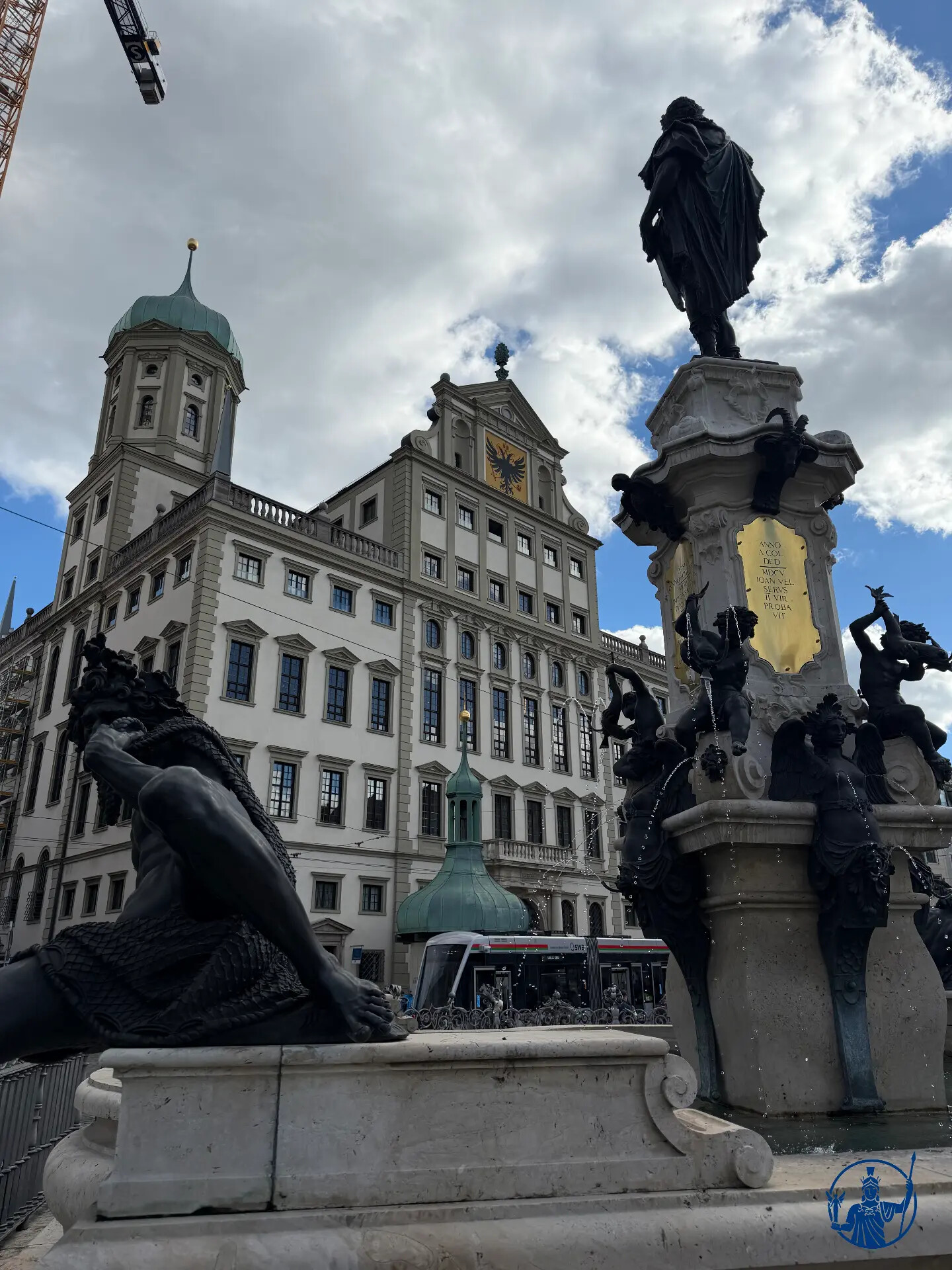 Augustusbrunnen auf dem Augsburger Rathausplatz mit Bronzefiguren und Brunnensäule, dahinter die Renaissancefassade des Rathauses und eine Straßenbahn