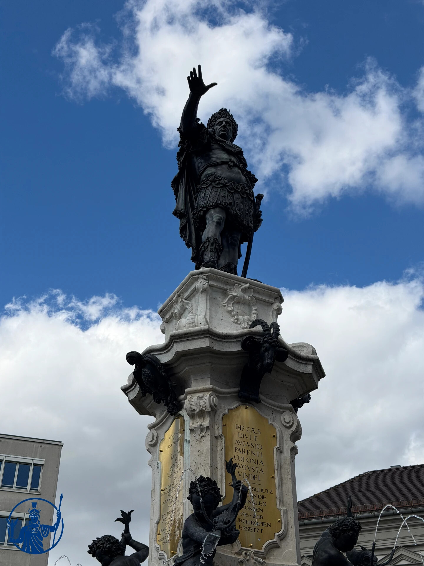 Bronzestatue des Kaisers Augustus auf der Säule des Augustusbrunnens vor blauem Himmel, darunter die vergoldete Inschriftentafel
