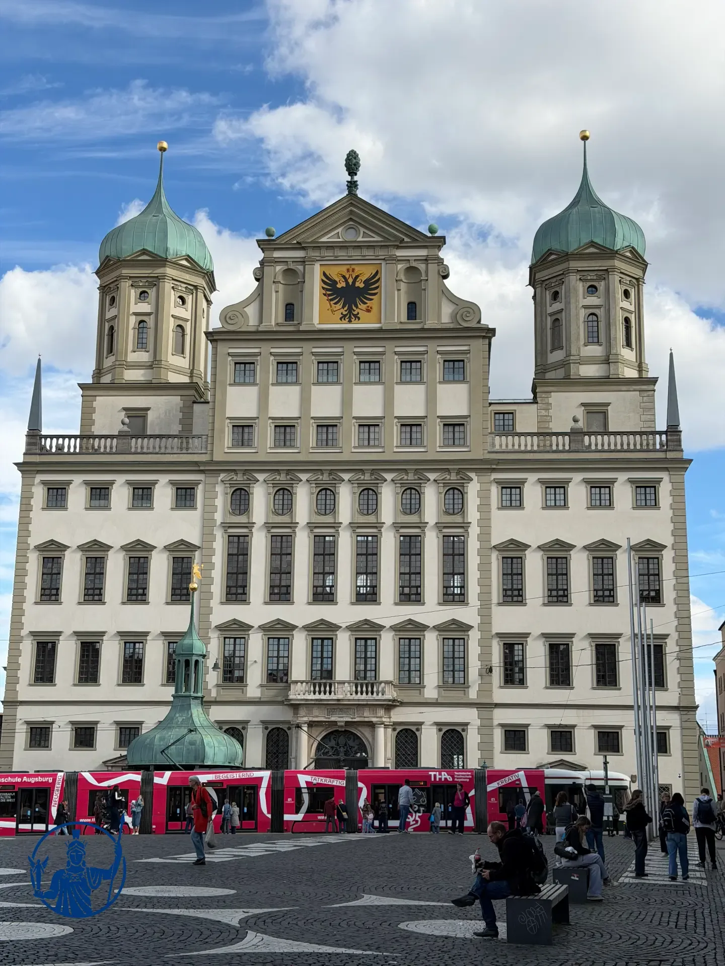 Frontalansicht des Augsburger Rathauses mit beiden Zwiebeltürmen und Reichsadlerwappen, davor eine rot-weiße Straßenbahn